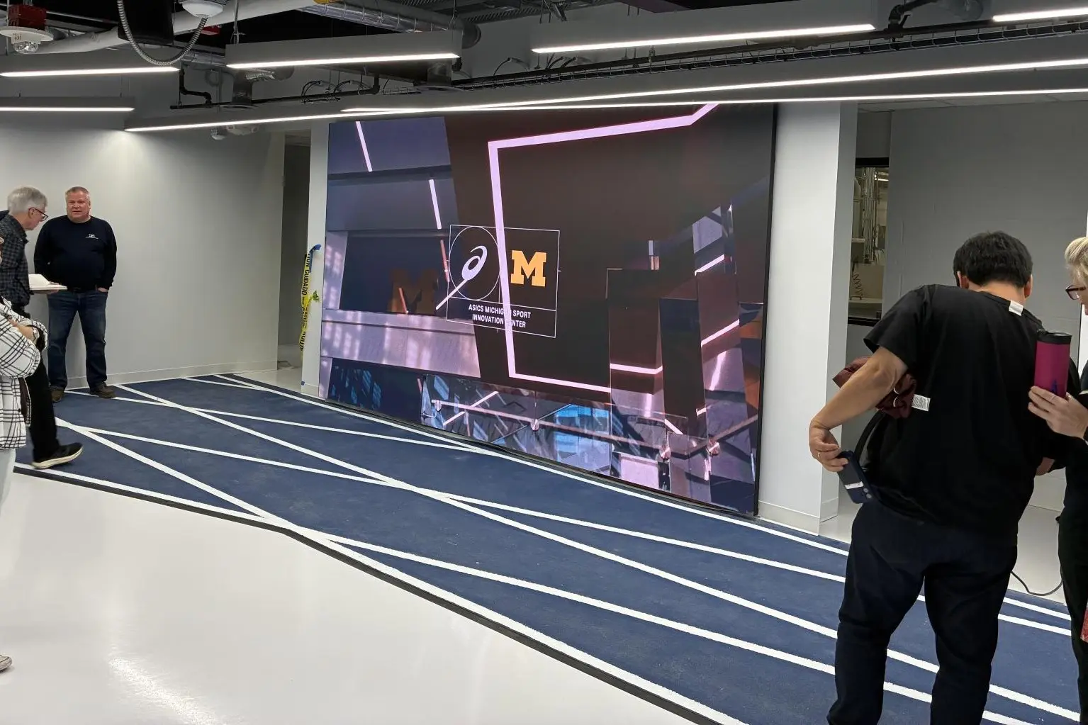 Large LED video wall at the end of an indoor running track, visitors looking on